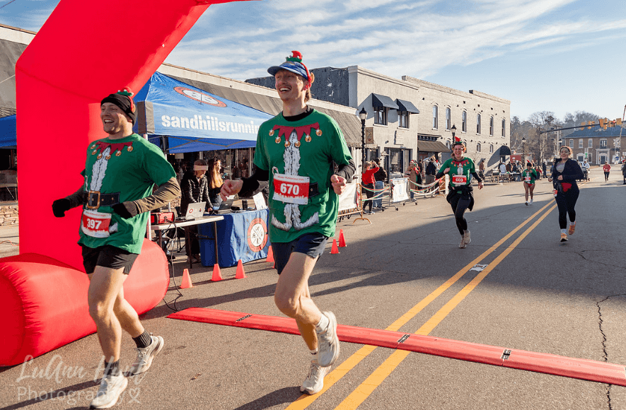 Two runners in matching green elf shirts cross the finish line side by side smiling
