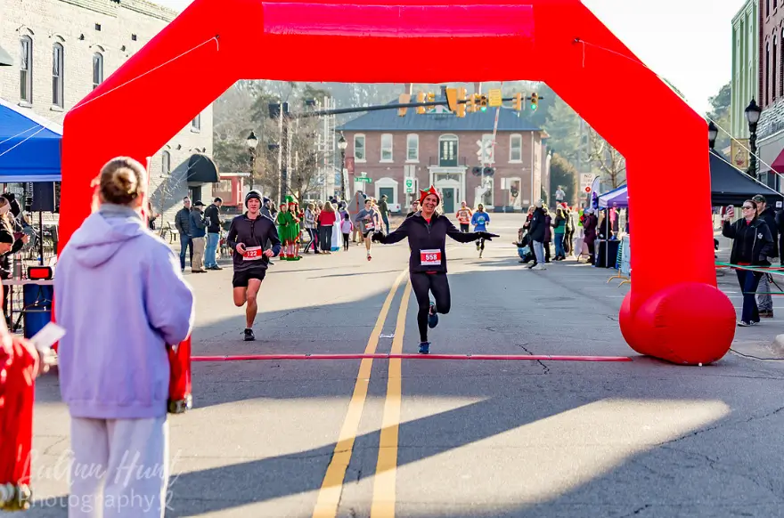 A runner in an elf hat crosses the finish line beneath a red inflatable arch with arms outstretched