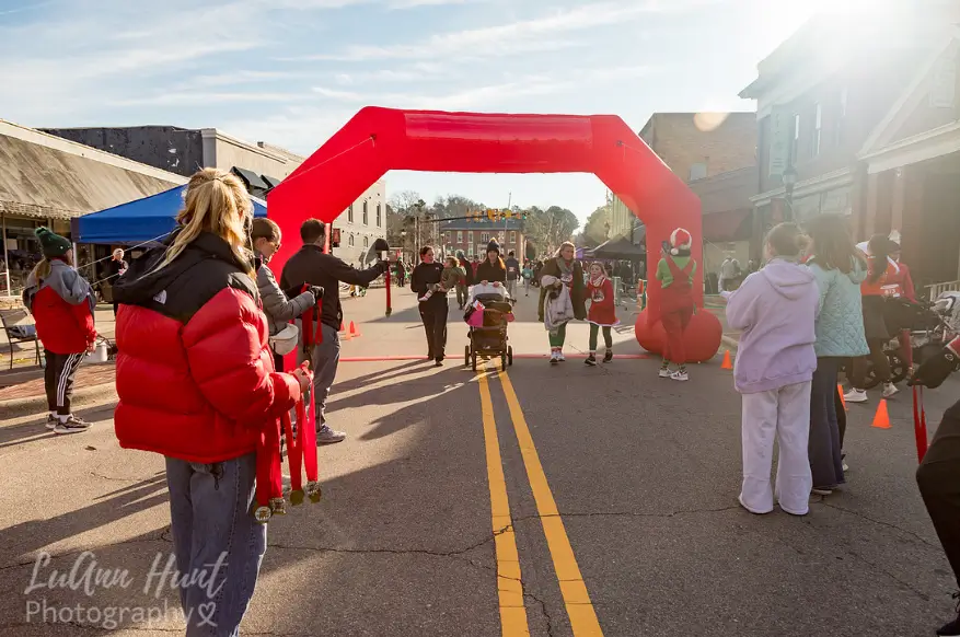 A volunteer in a red jacket holds finisher medals as runners pass under the arch in morning light