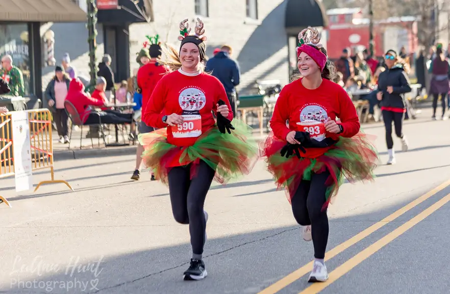 Two runners in red holiday shirts, tulle tutus and reindeer antlers run side by side