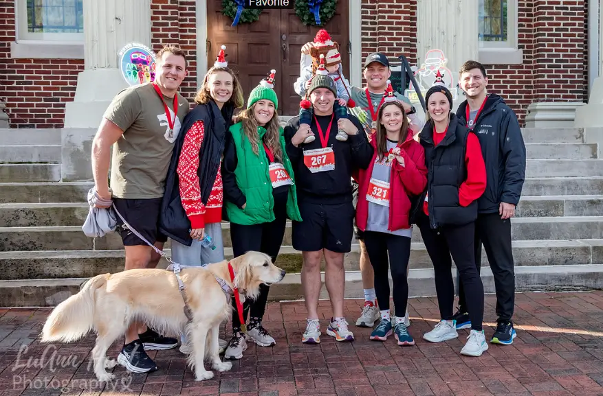 A group of runners and a dog wearing a finisher medal pose on brick steps in front of a wreathed door